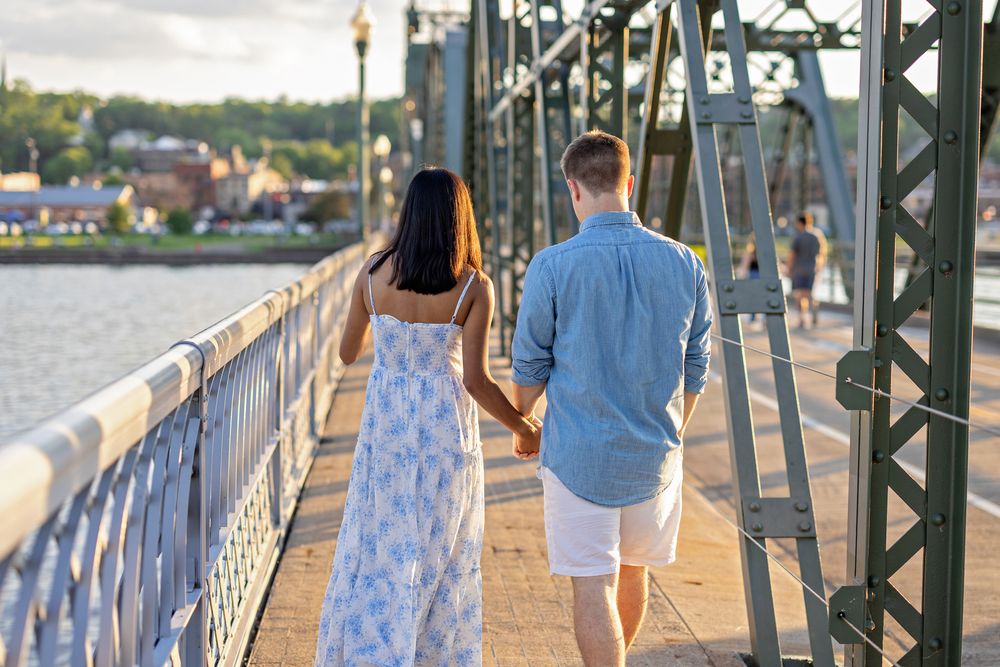 Couple walking hand in hand on a green metal bridge with a river and town visible behind them.