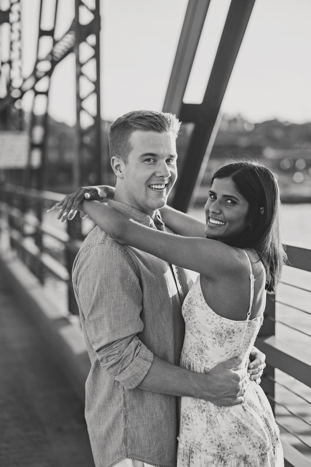 Black and white photo of a smiling couple hugging on a bridge at sunset.