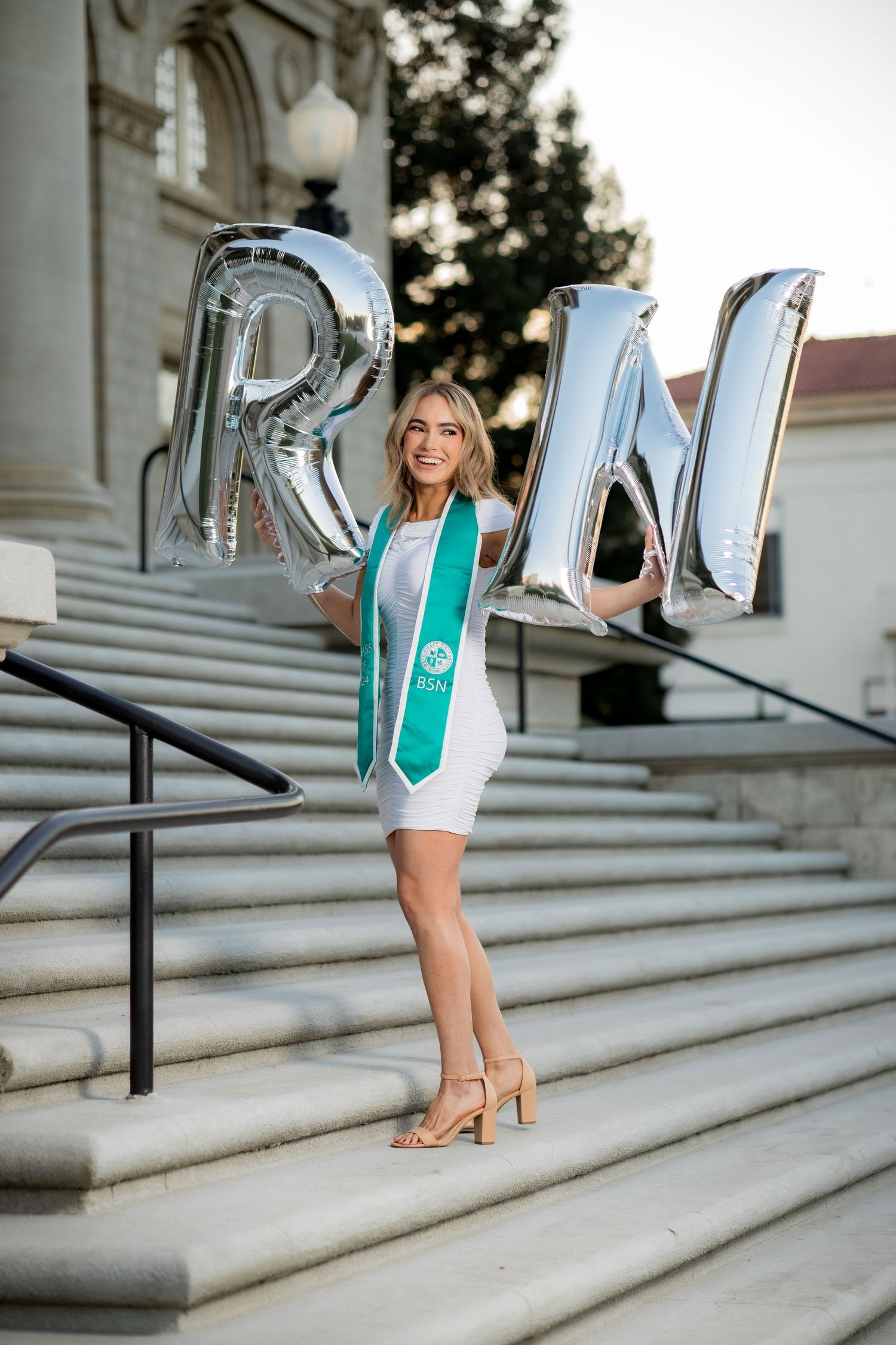 Blonde graduate in white dress holding silver RN balloon letters on university steps.