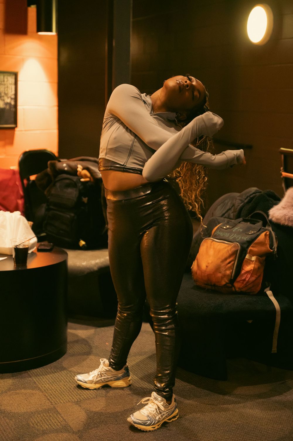A woman in metallic leggings and sneakers strikes a dramatic pose in a dimly lit lounge area.