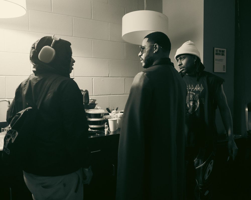 Three men in casual streetwear having a conversation in a black and white indoor setting.