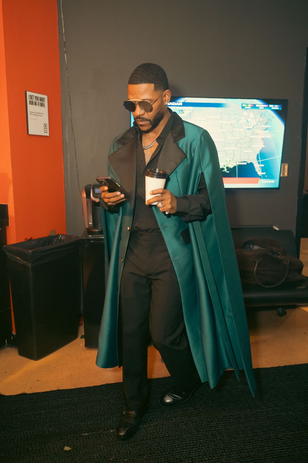 Stylish man in teal cape coat and sunglasses checks phone backstage, holding a cup near a weather TV screen.