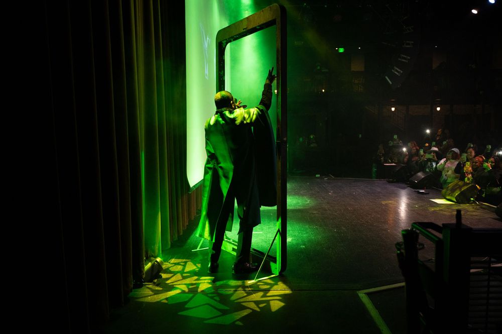 Performer on stage bathed in dramatic green light, standing in a doorframe during a theatrical production.