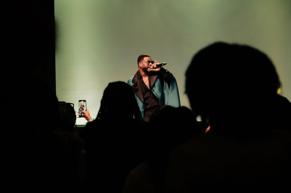 A male performer sings into a microphone on a dimly lit stage while audience members record with smartphones.