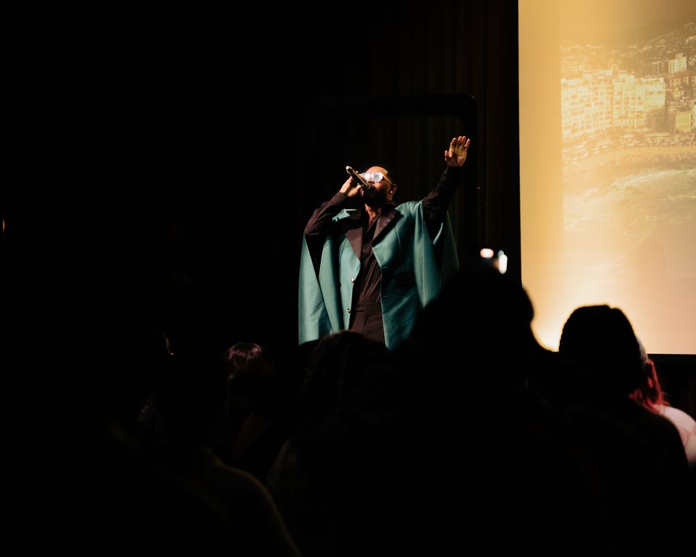 A performer in a teal suit sings into a microphone on a dimly lit stage before a crowd.