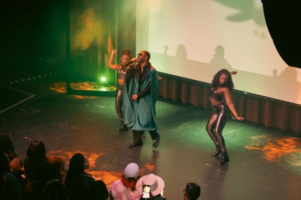 Three performers on stage, with two dancers in metallic outfits flanking a singer under green and orange stage lighting.