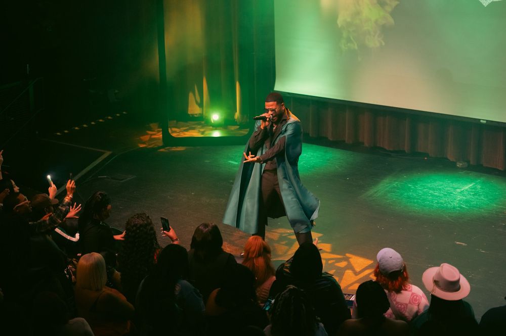 A performer sings on a green-lit stage while an enthusiastic crowd reaches out and records with phones.