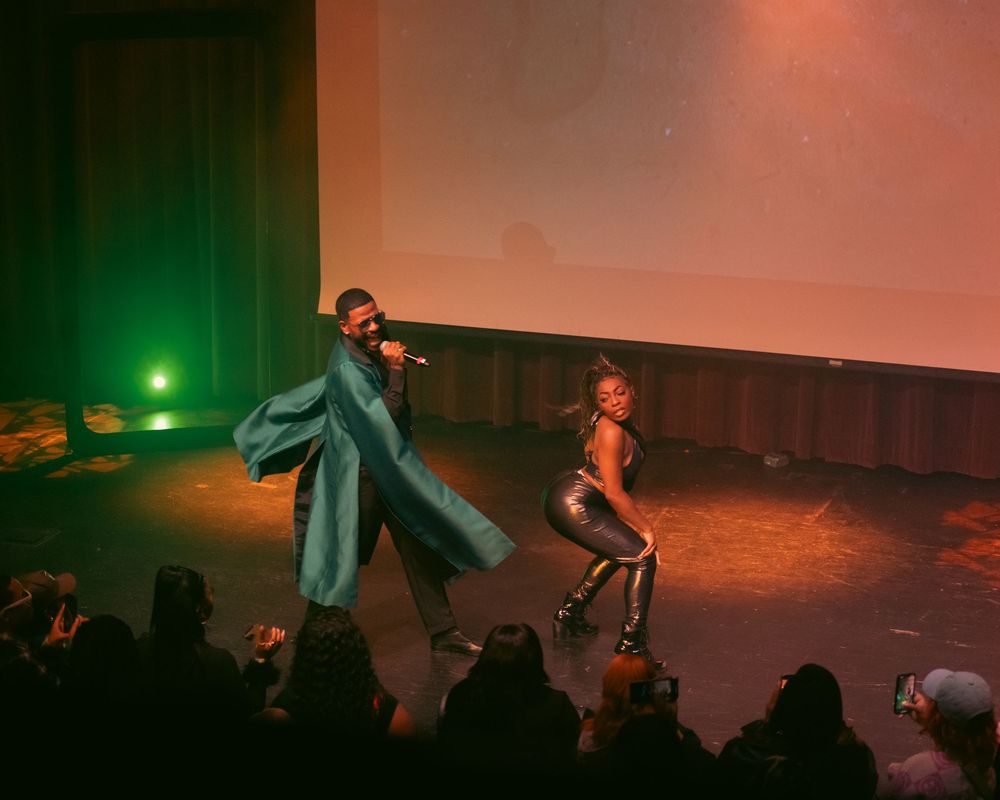 Two performers on stage, one in a teal coat with a mic, another in silver outfit dancing under green lights.