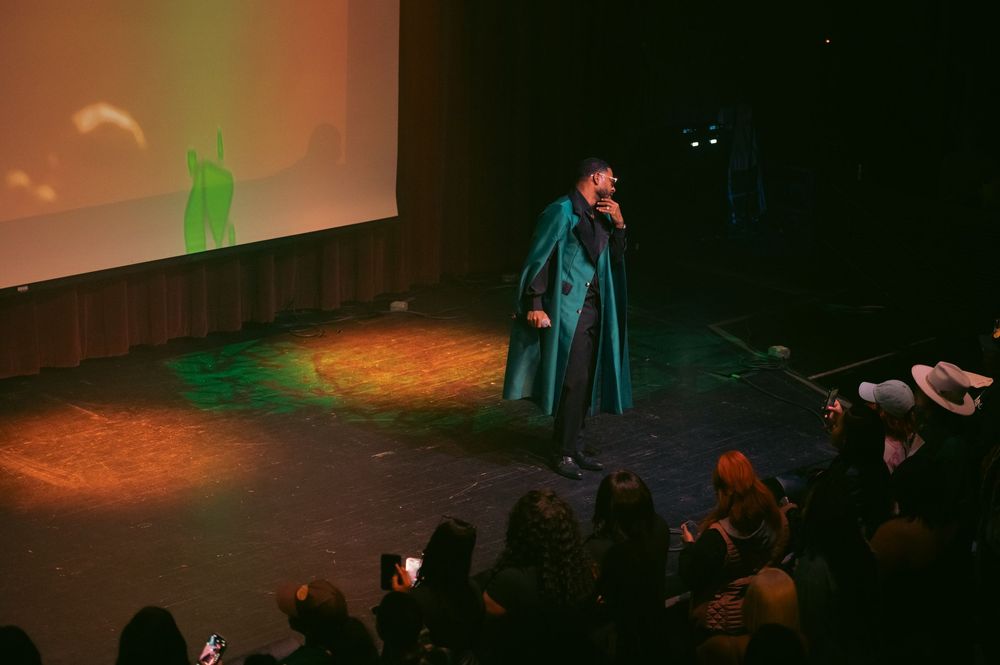 Performer in teal cape sings on stage with green projected backdrop and audience silhouettes in foreground.
