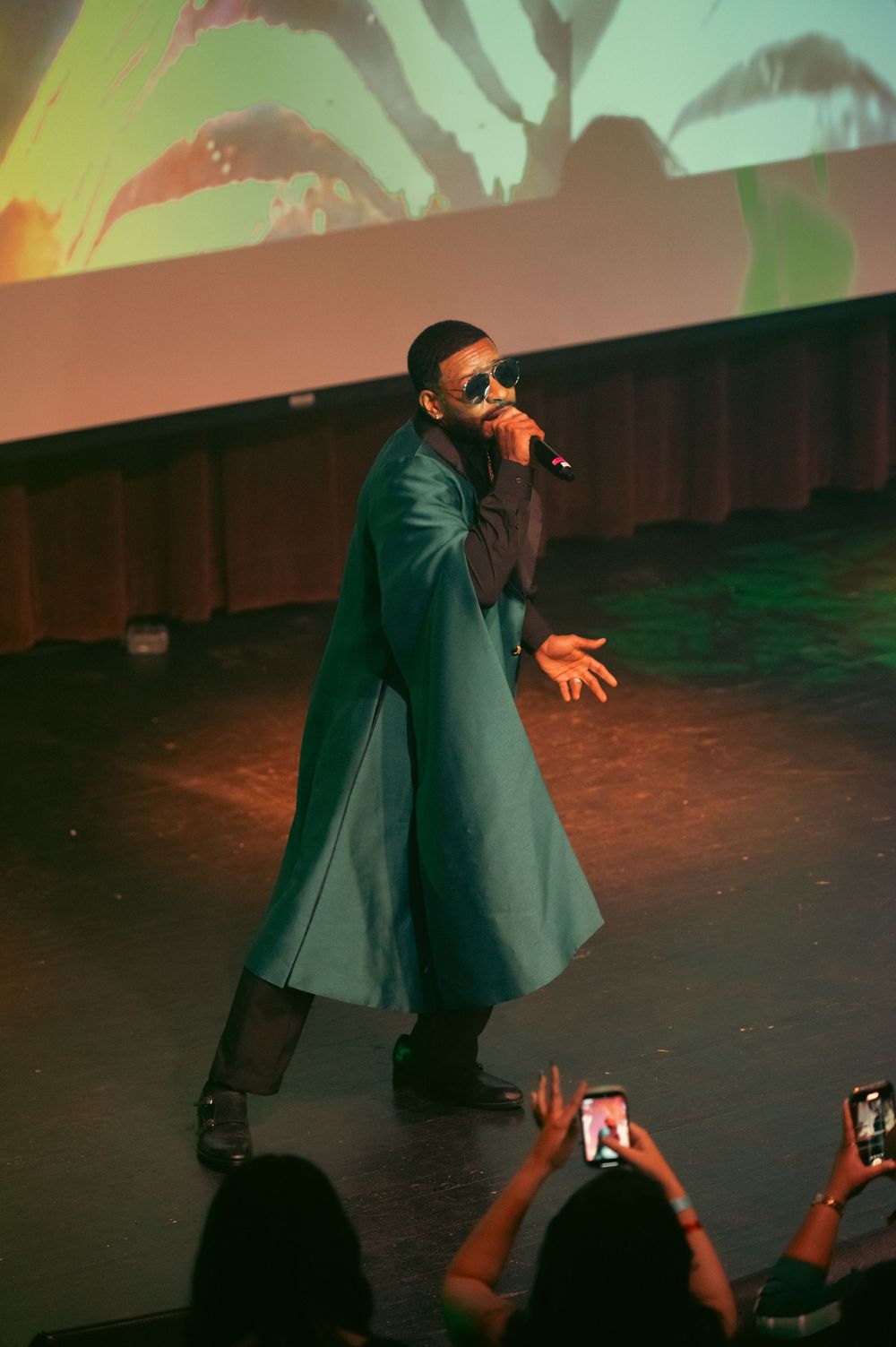 A performer in a teal trench coat sings into a microphone on stage with a colorful projection backdrop.