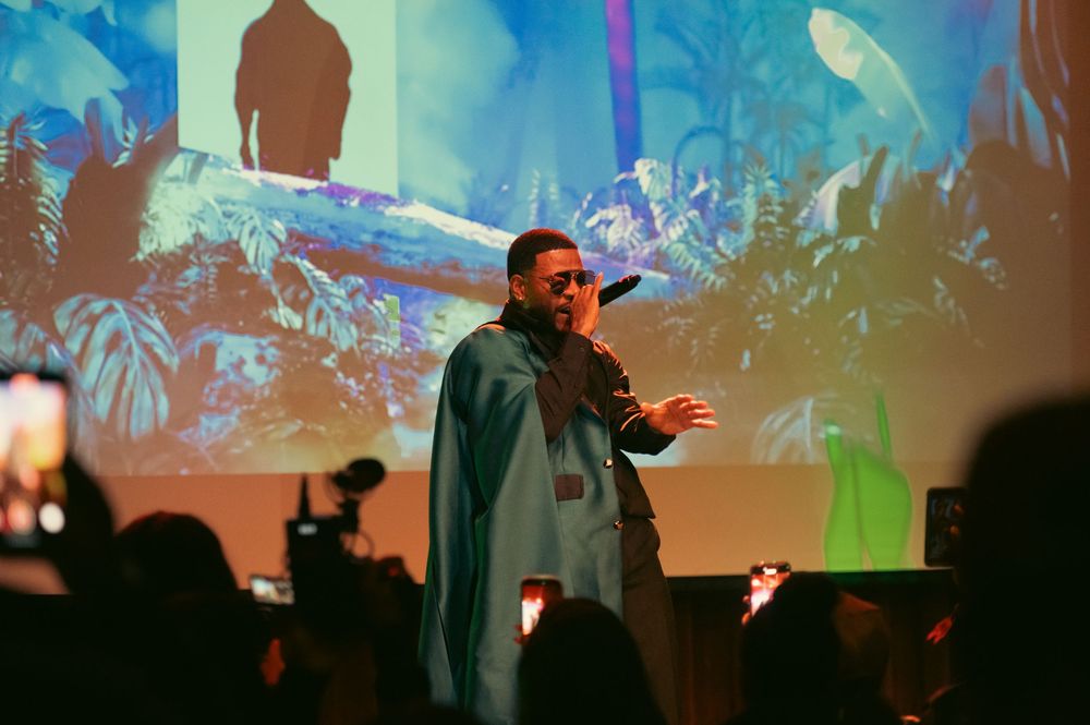 A performer in a blue cape raps on stage with a tropical projection backdrop at an indoor concert event.
