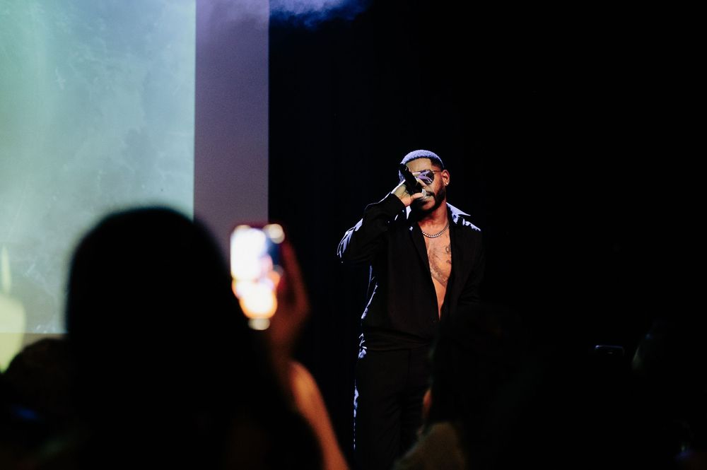Male performer singing on dark stage wearing black jacket and cap, with audience member filming on smartphone in foreground.