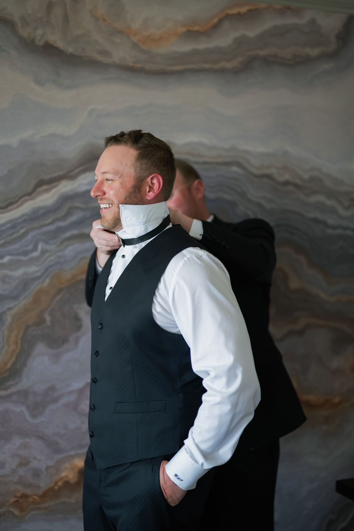 Groom smiling as groomsman helps adjust his bow tie before wedding ceremony against marble wall backdrop.