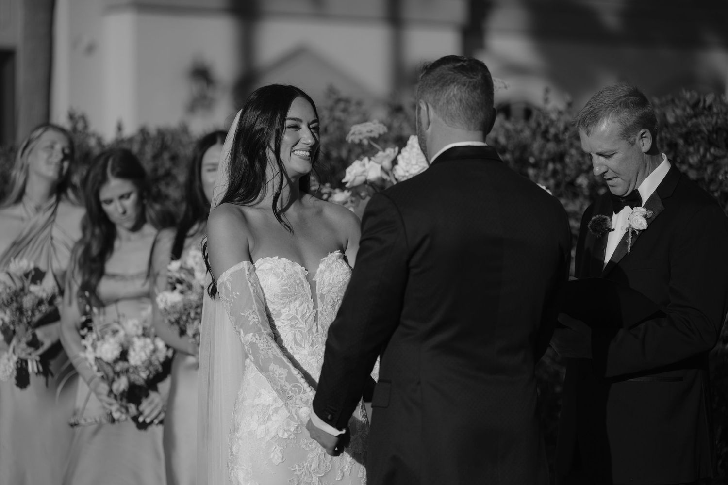 Black and white photo of a bride and groom holding hands during an outdoor wedding ceremony with bridesmaids in background.