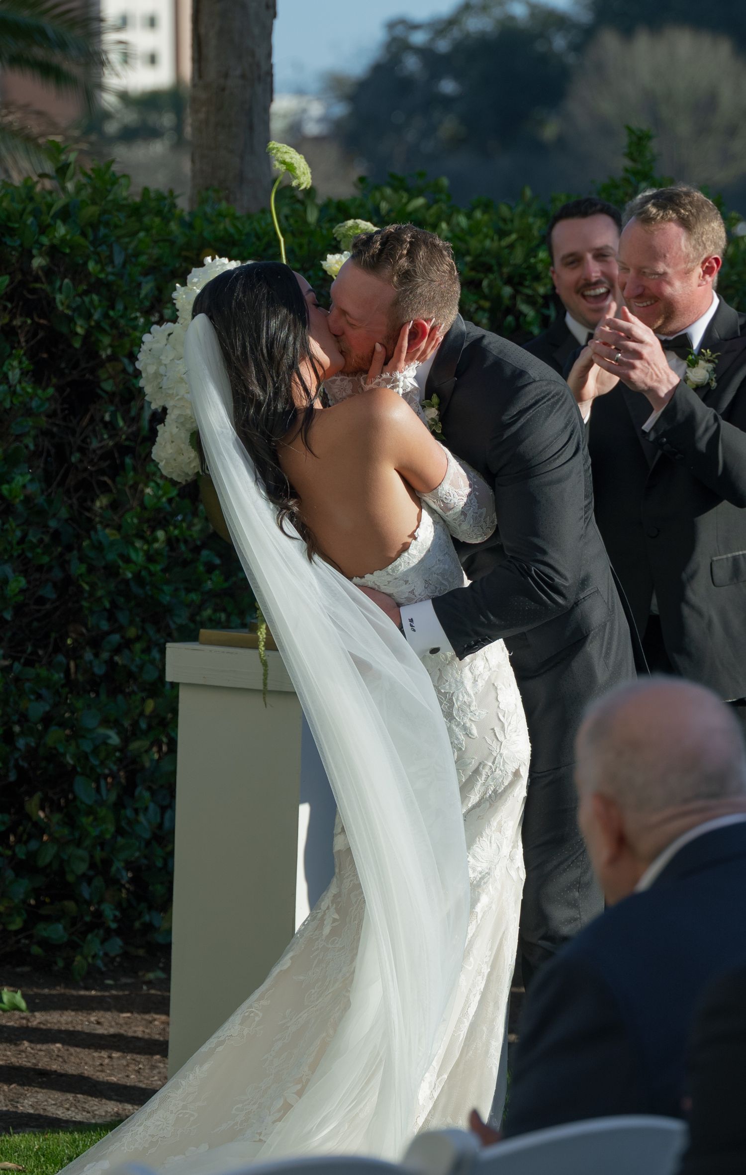 Bride and groom share first kiss at outdoor wedding ceremony as groomsmen laugh and celebrate behind them.