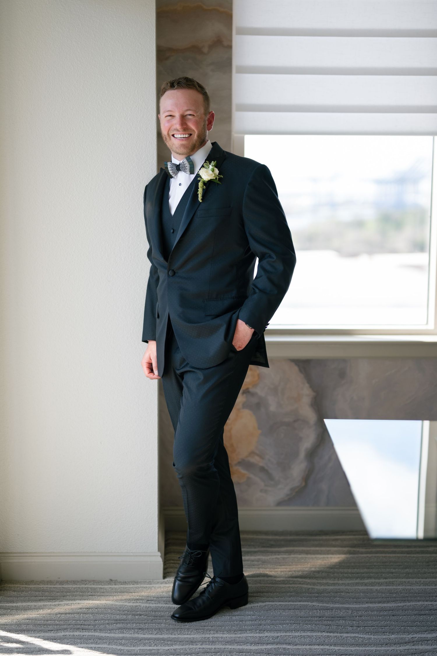 Dapper groom in black three-piece tuxedo with boutonniere leaning against window wall smiling.