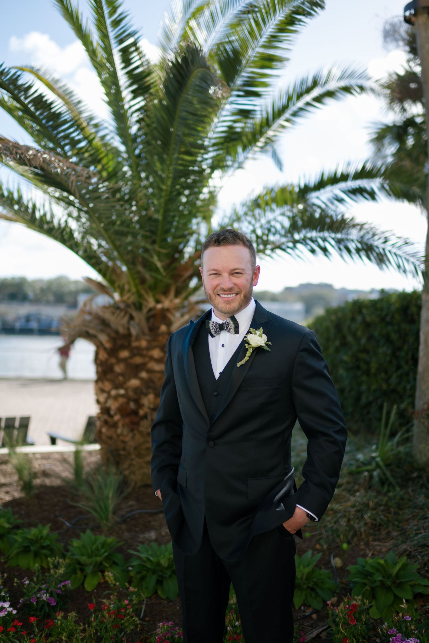 Smiling groom in black tuxedo with boutonniere stands before a tropical palm tree at an outdoor wedding venue.
