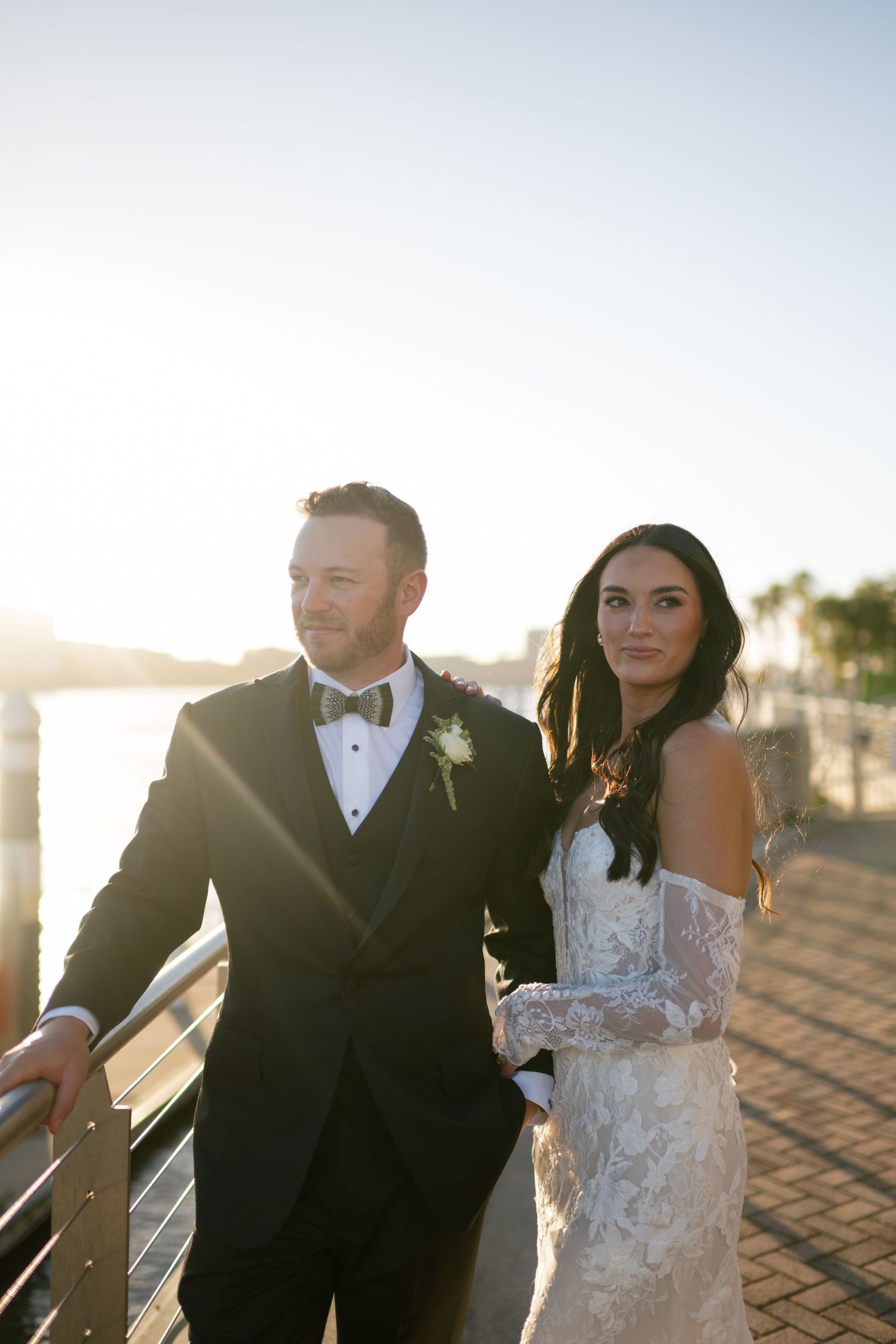 Bride and groom posing at sunset on a waterfront promenade, she wearing lace off-shoulder gown, he in black tuxedo.