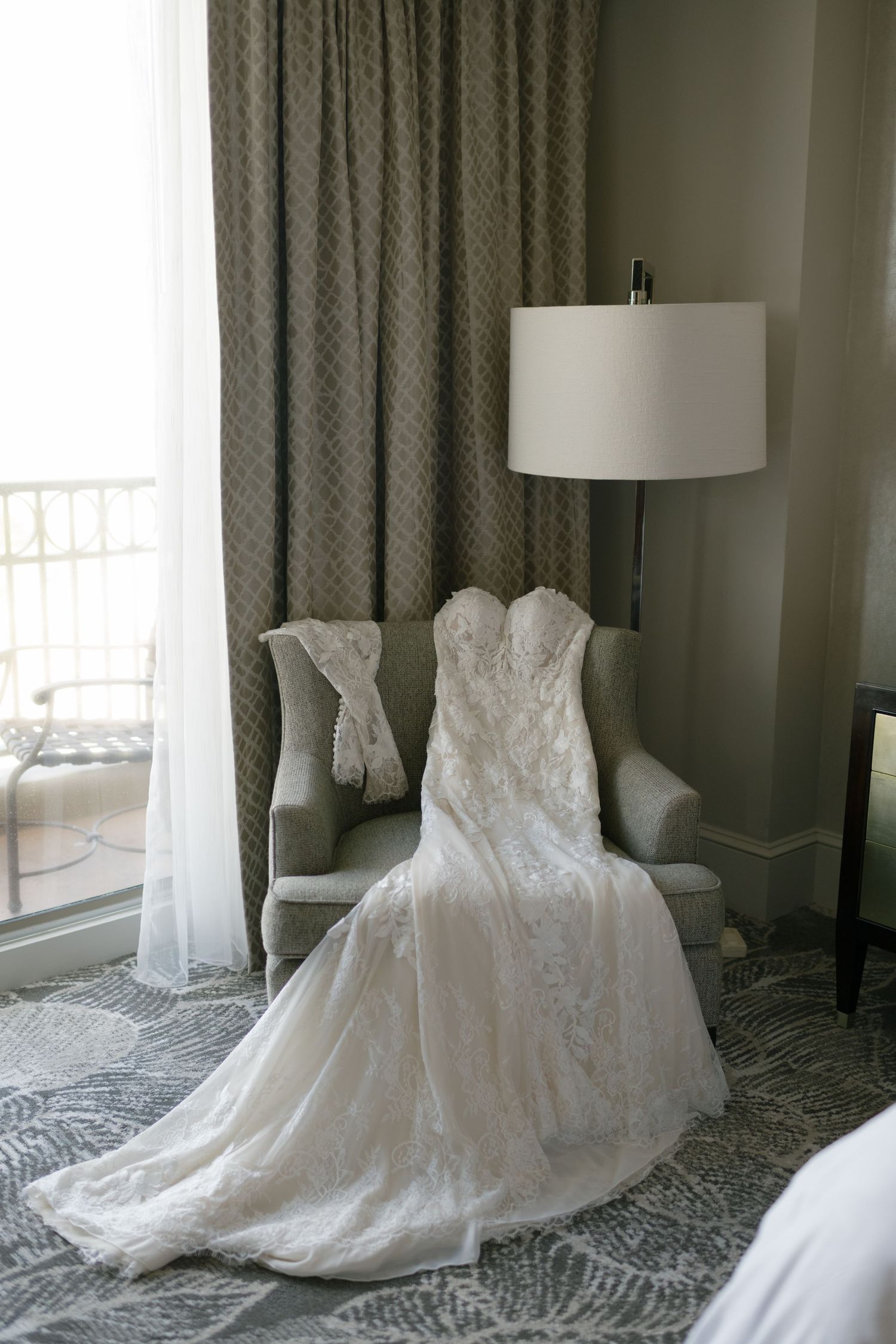 Elegant white lace strapless wedding gown draped over a gray armchair beside a lamp in a hotel room.