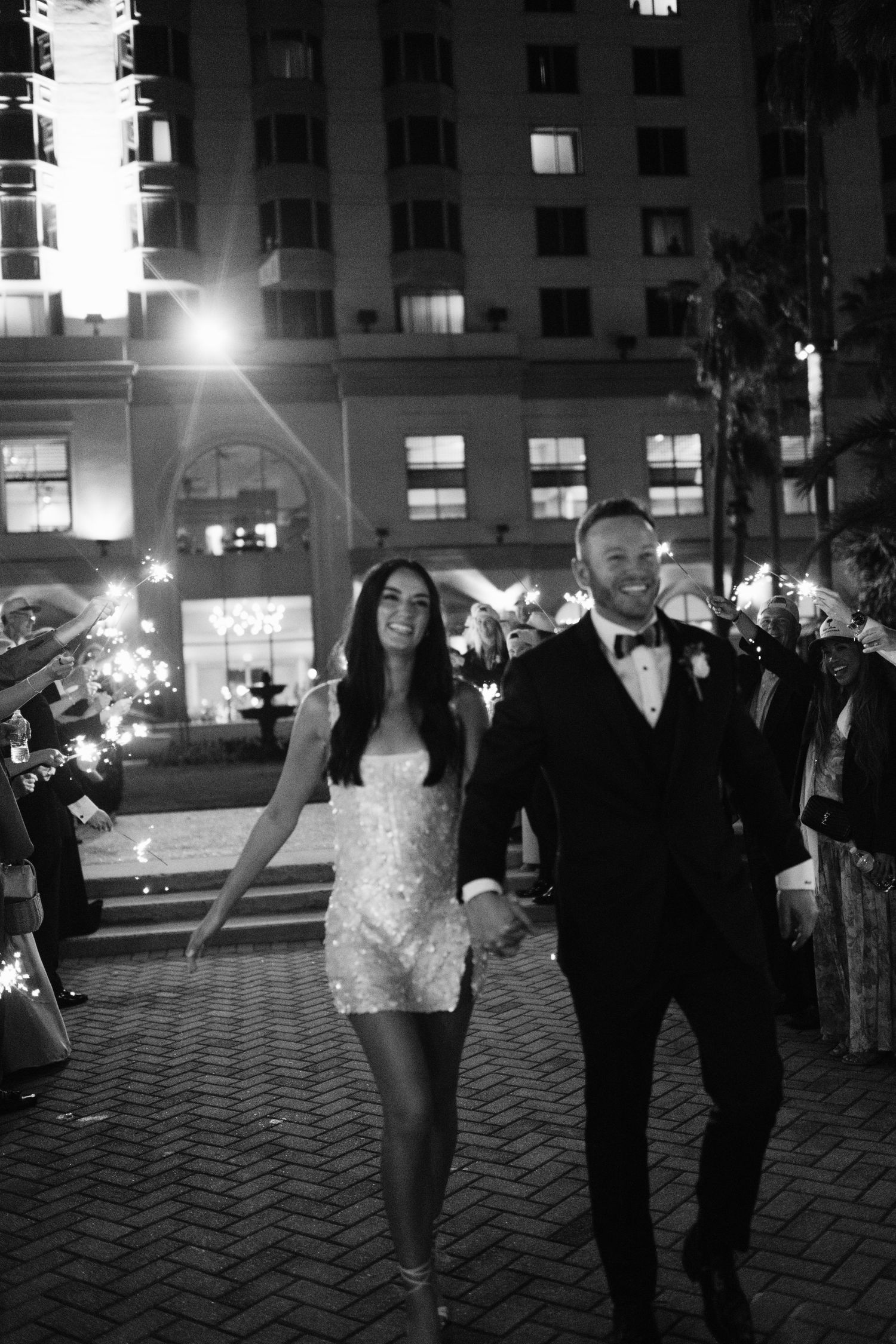Black and white photo of a bride and groom holding hands walking through a sparkler exit at their wedding reception.