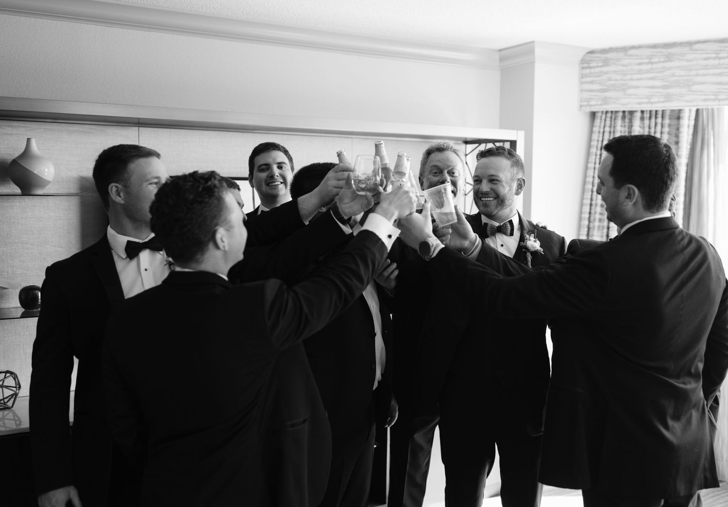 Black and white photo of groomsmen in tuxedos toasting with glasses during a wedding celebration indoors.