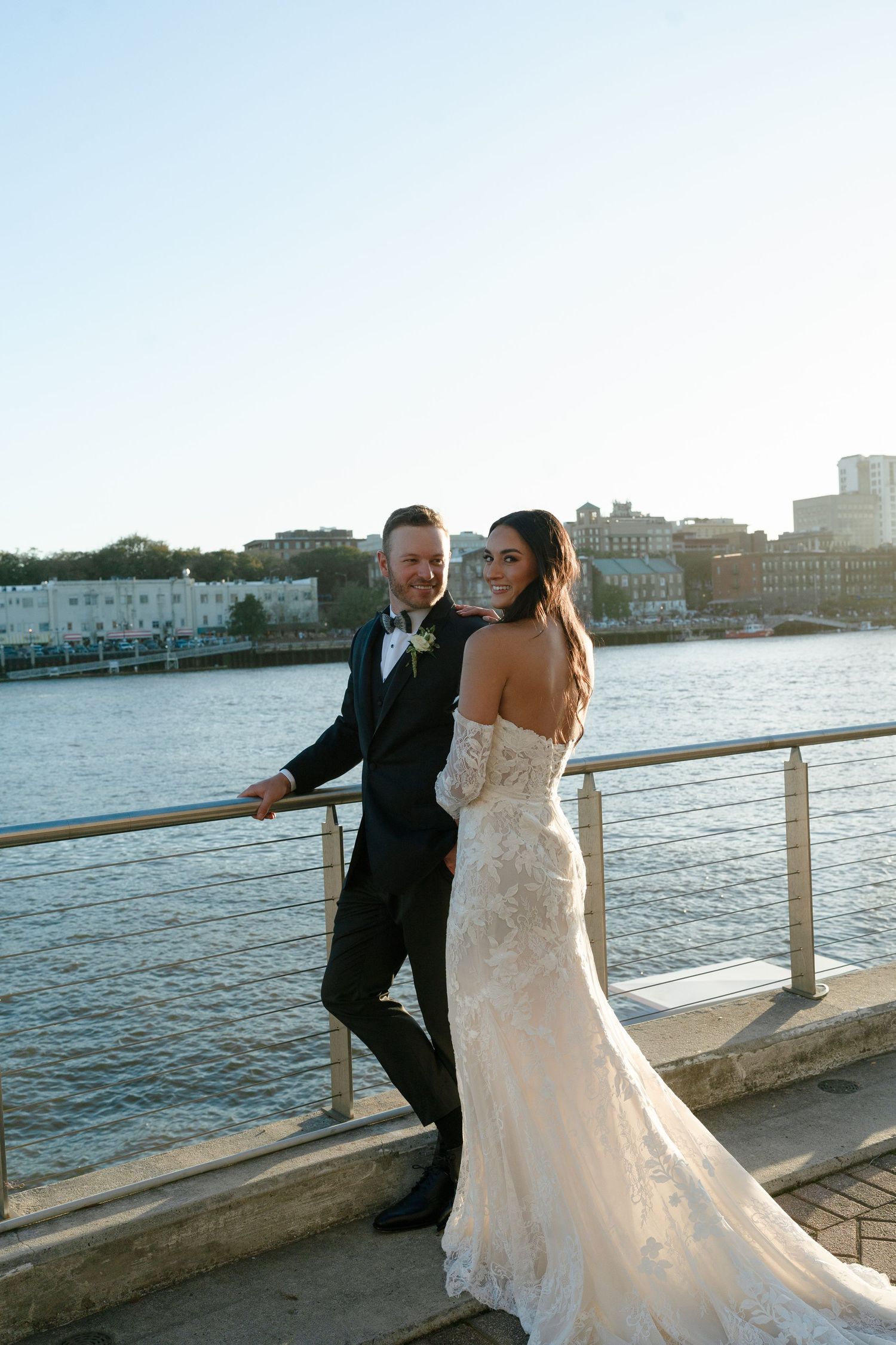 Bride and groom posing on a waterfront dock in Savannah, GA, wearing a lace gown and black tuxedo.