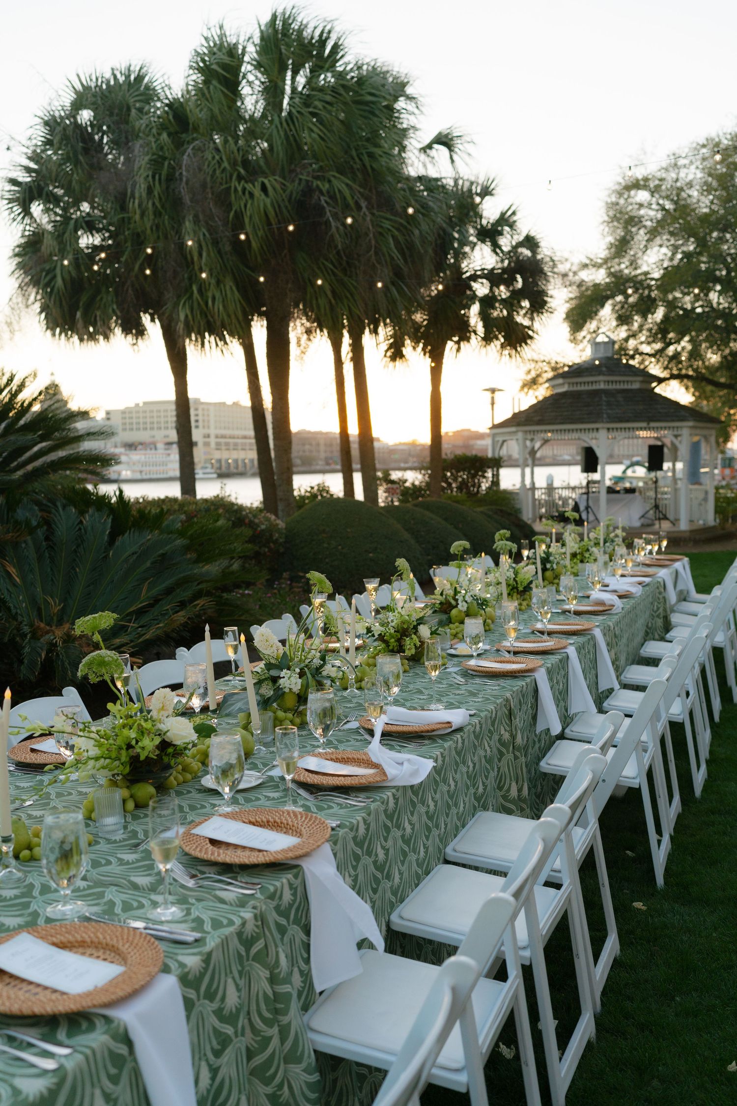 Elegant outdoor wedding reception with long table, green floral linens, white chairs, palm trees, and gazebo at sunset.
