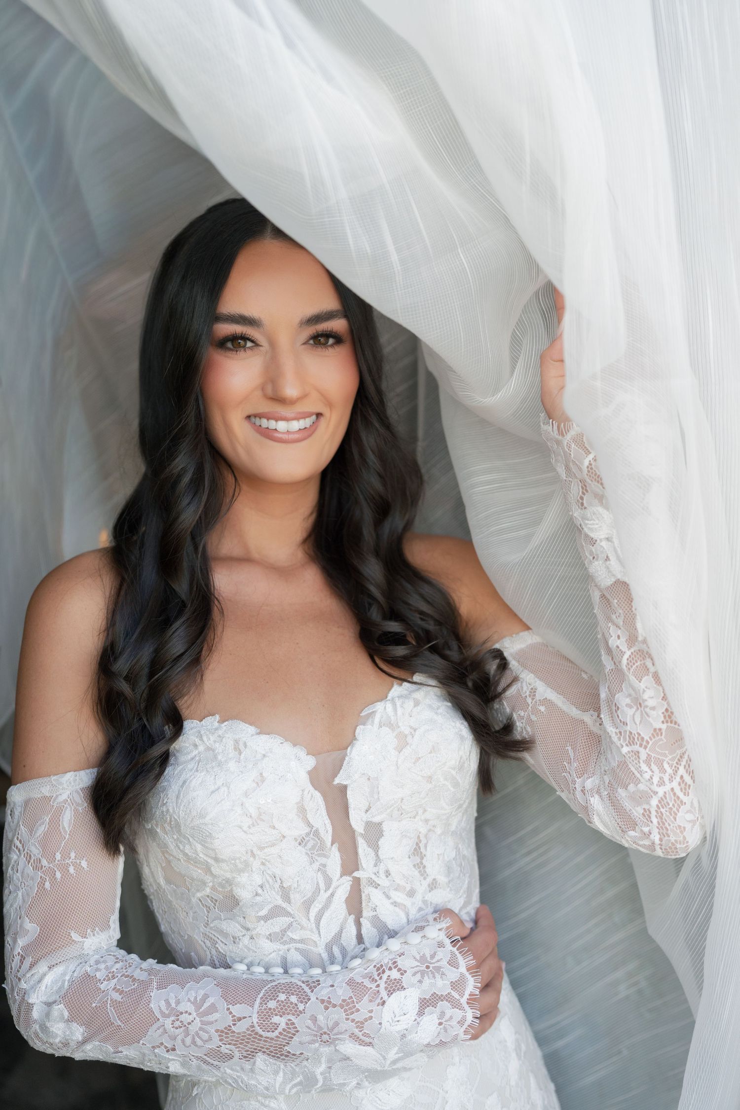 Smiling bride in lace wedding gown with sheer sleeves, holding white tulle veil, long dark wavy hair.