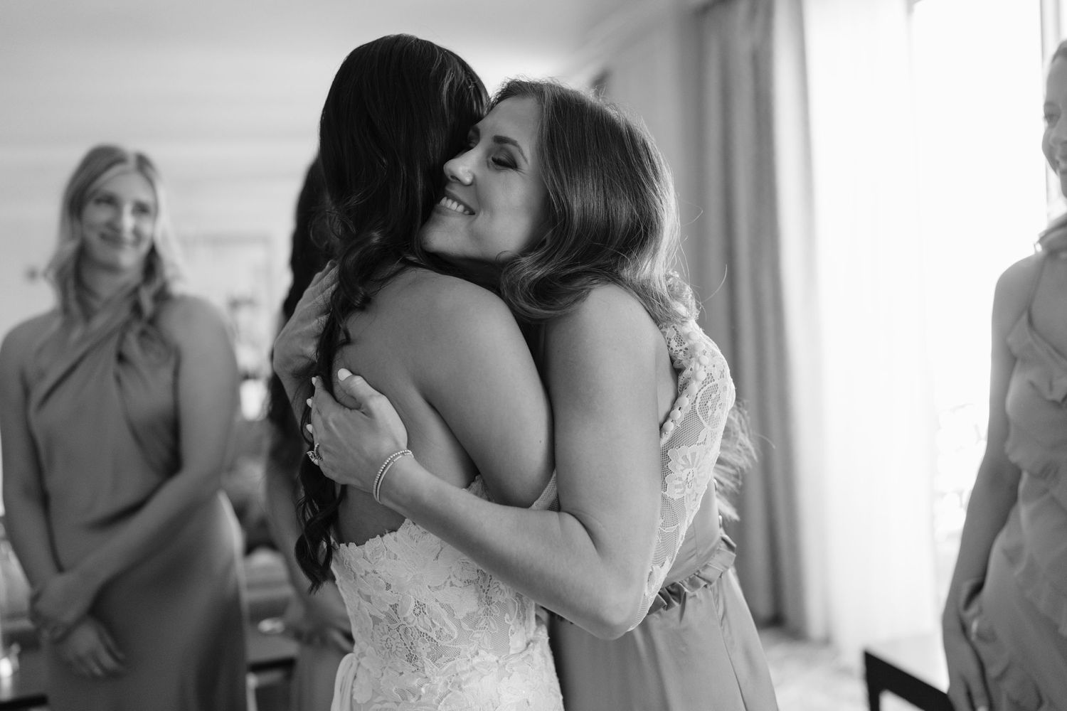 Bride in lace gown shares heartfelt hug with bridesmaid during wedding preparations in black and white photo.