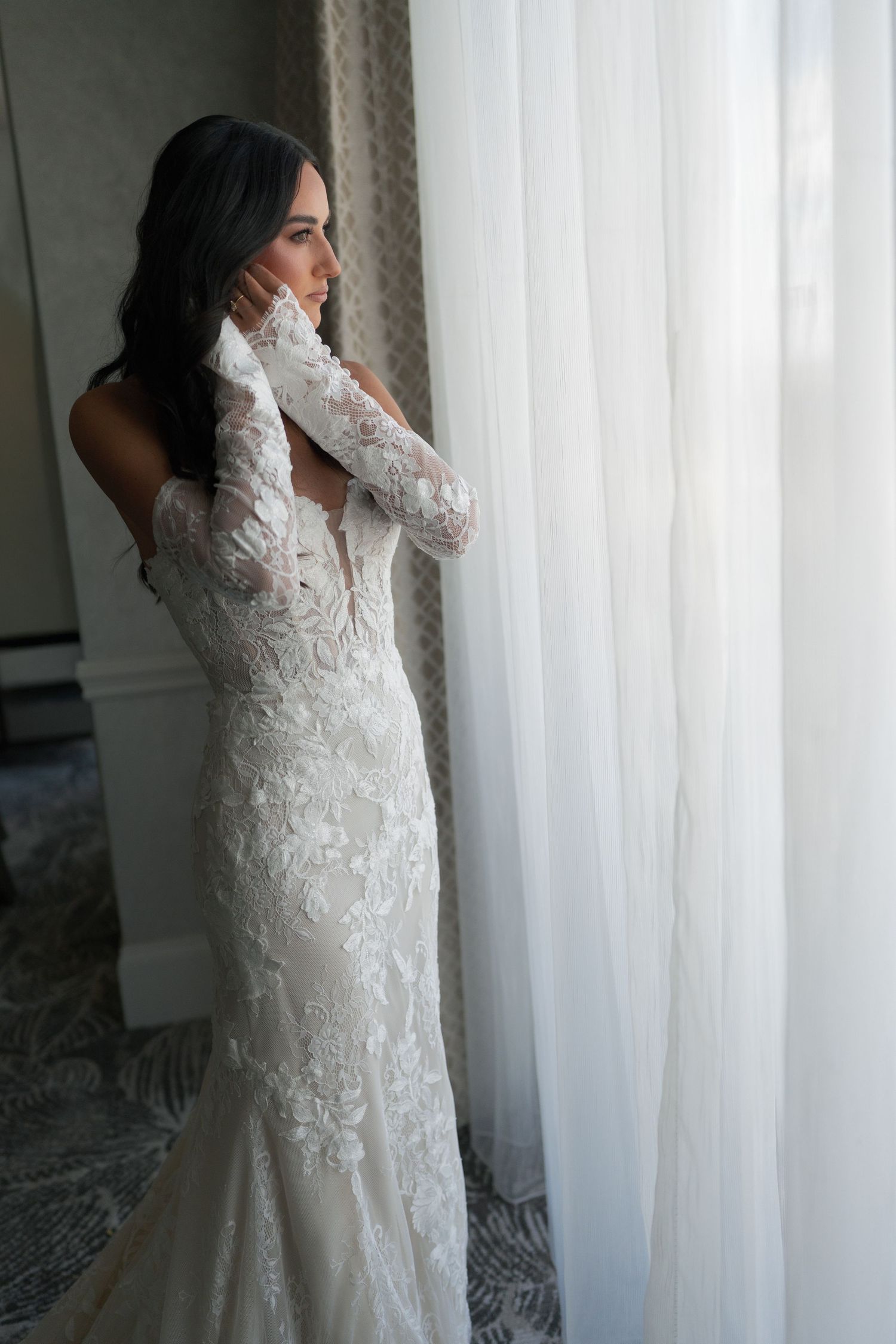 Elegant bride in a lace long-sleeve wedding gown gazing out a window in soft natural light.