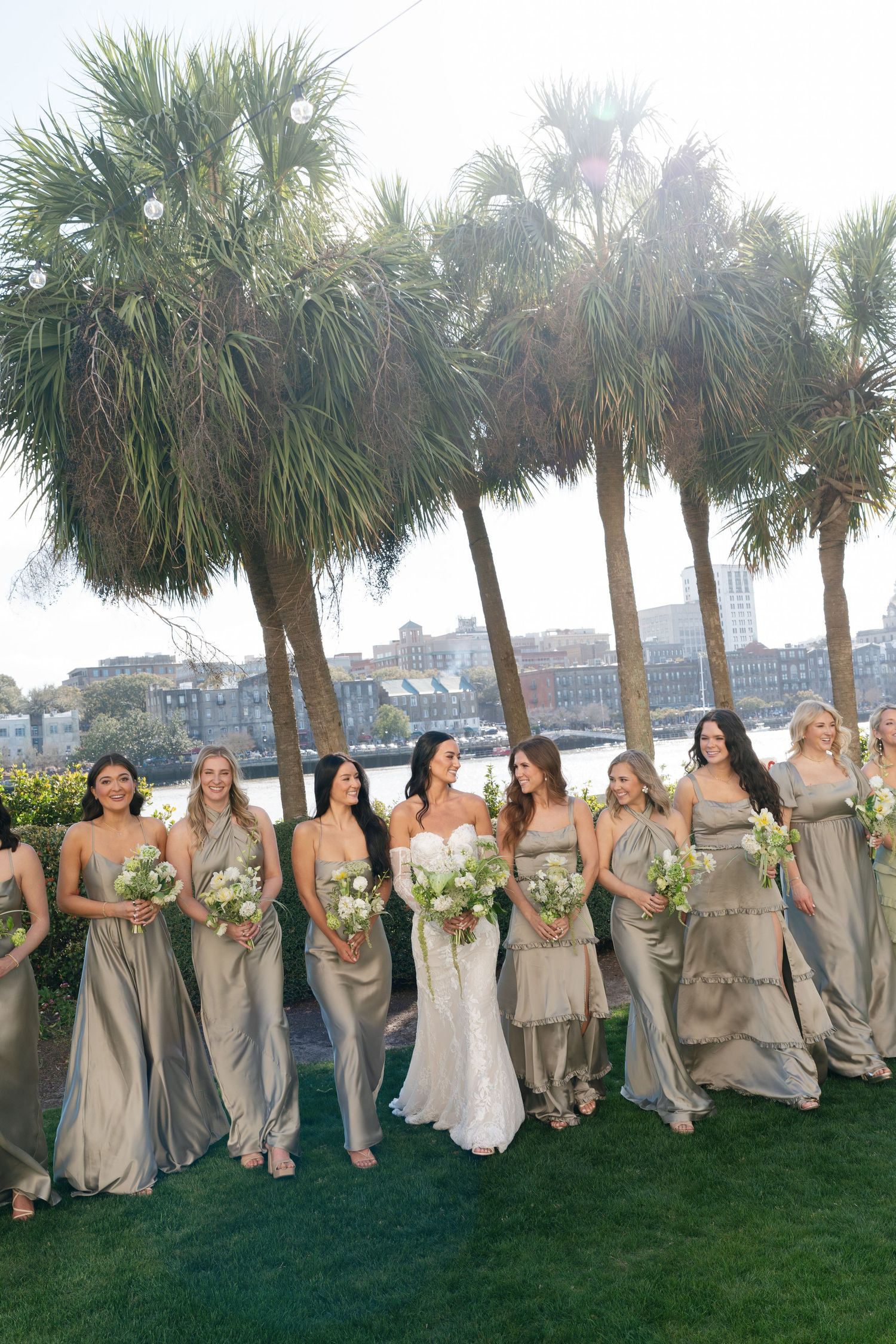 Bride with bridesmaids in champagne satin gowns holding white bouquets near palm trees waterfront.