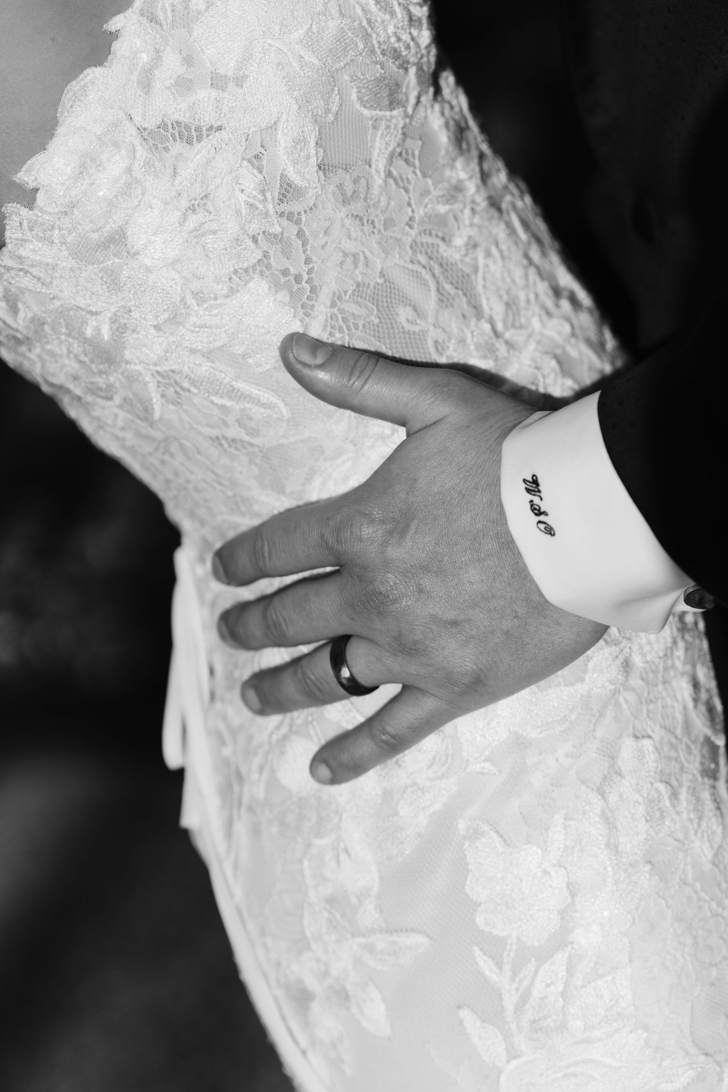 Close-up black and white photo of groom's hand with wedding ring resting on bride's lace wedding dress.