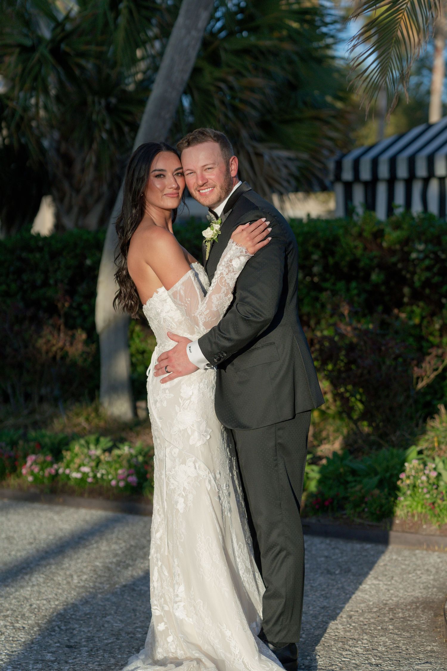 Bride in lace gown and groom in dark suit embrace outdoors at tropical wedding venue surrounded by palm trees.