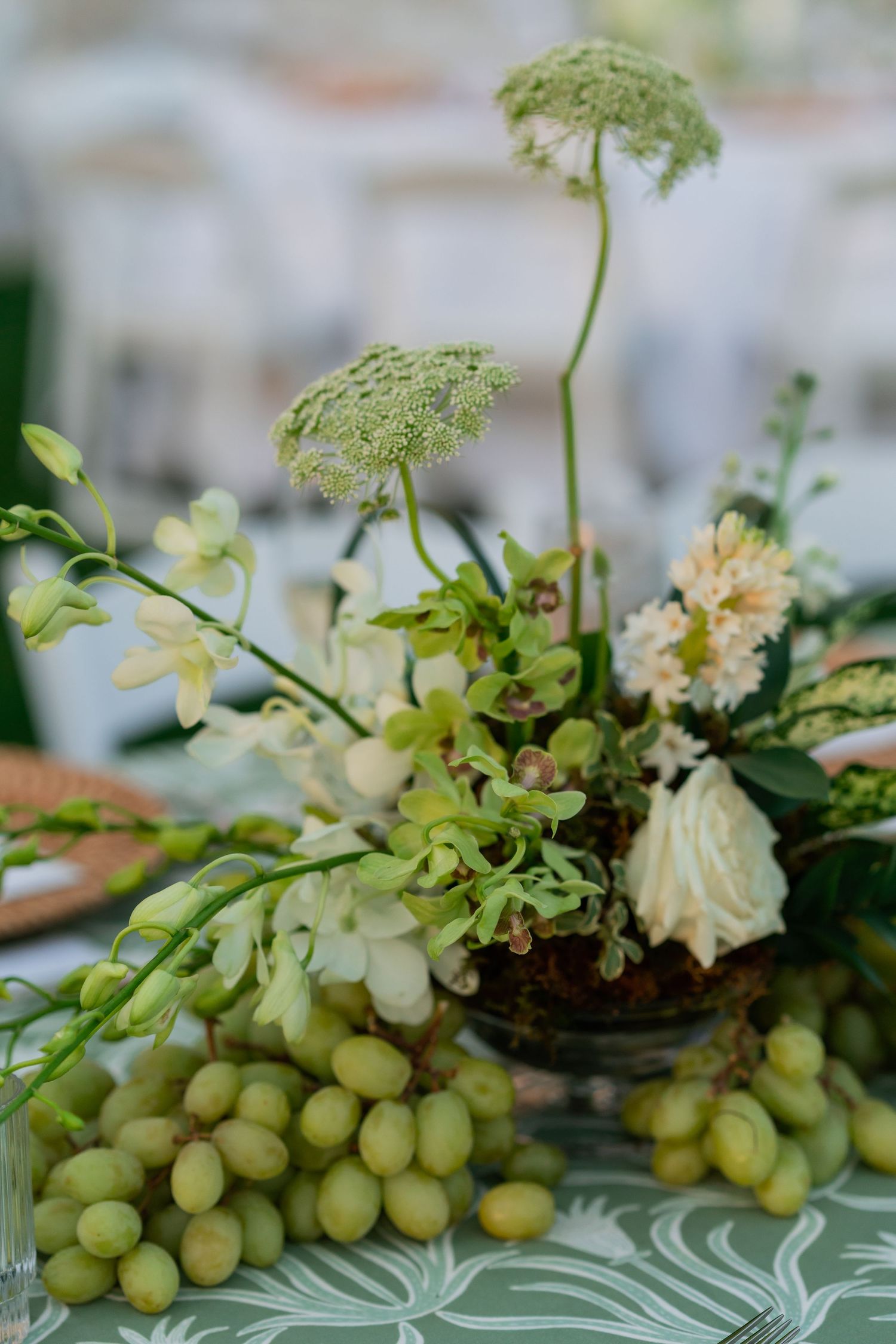Elegant wedding centerpiece with green orchids, white roses, and fresh grapes on a floral tablecloth.