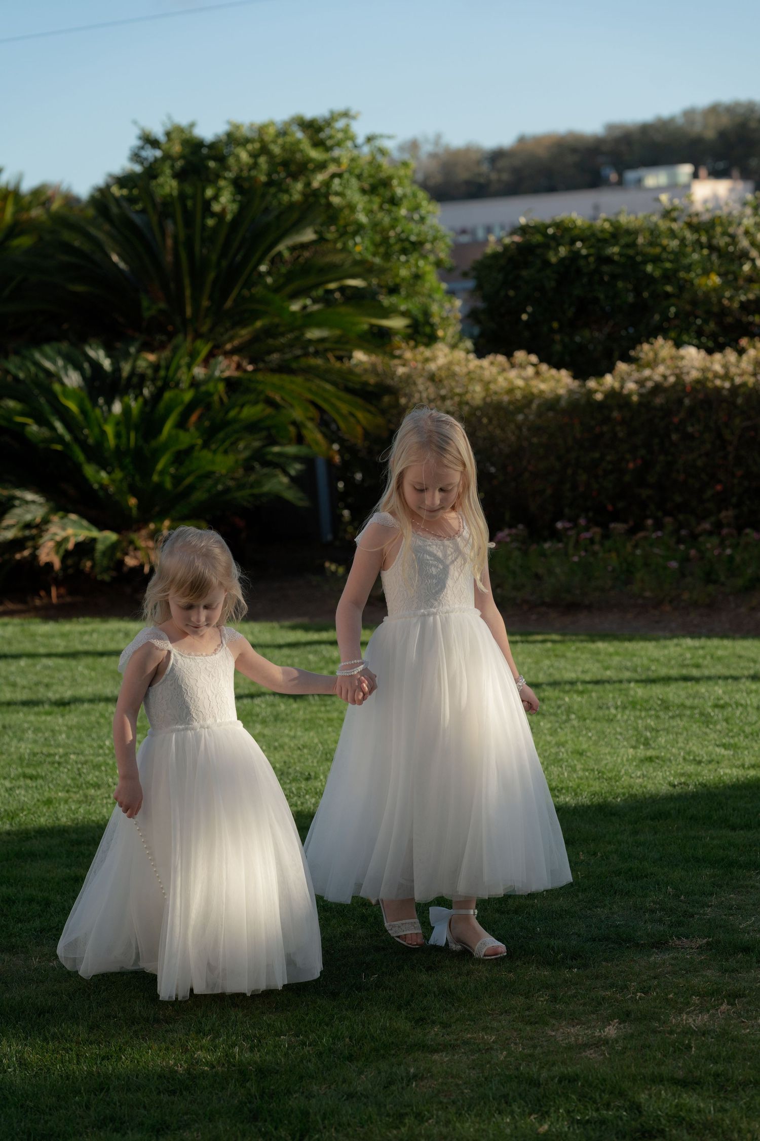 Two young flower girls in white tulle dresses holding hands walking on green lawn at wedding.