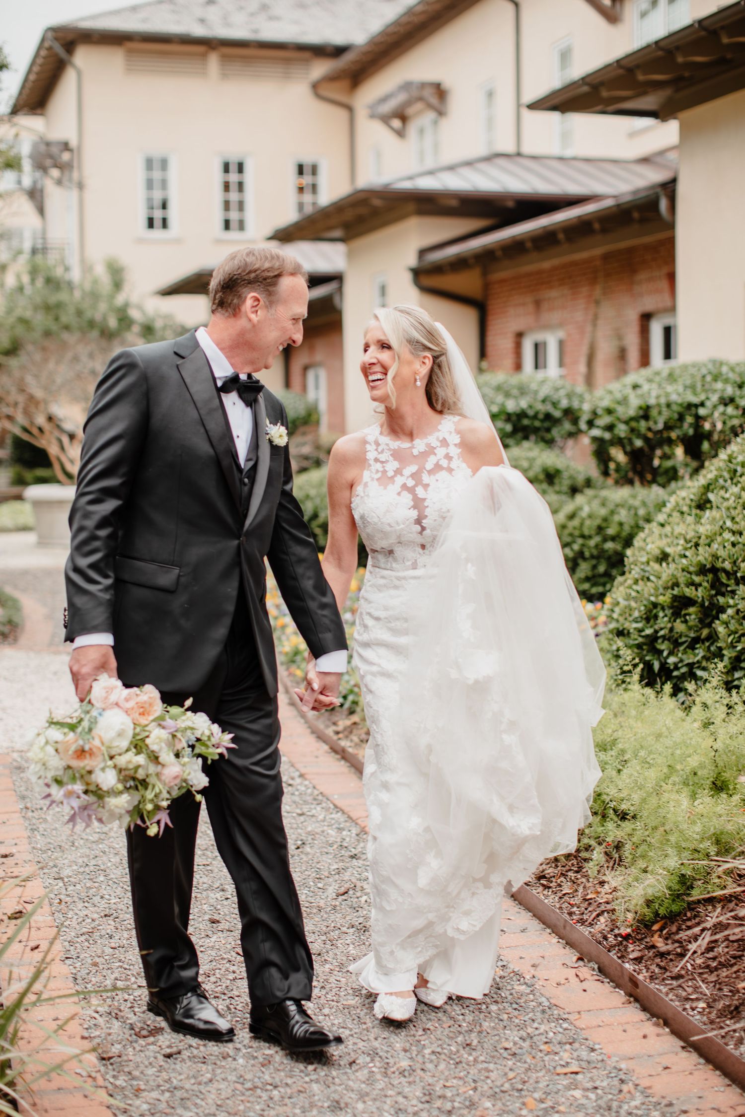 Bride and groom walking hand in hand, laughing joyfully outside an elegant venue, bride in lace gown with veil.