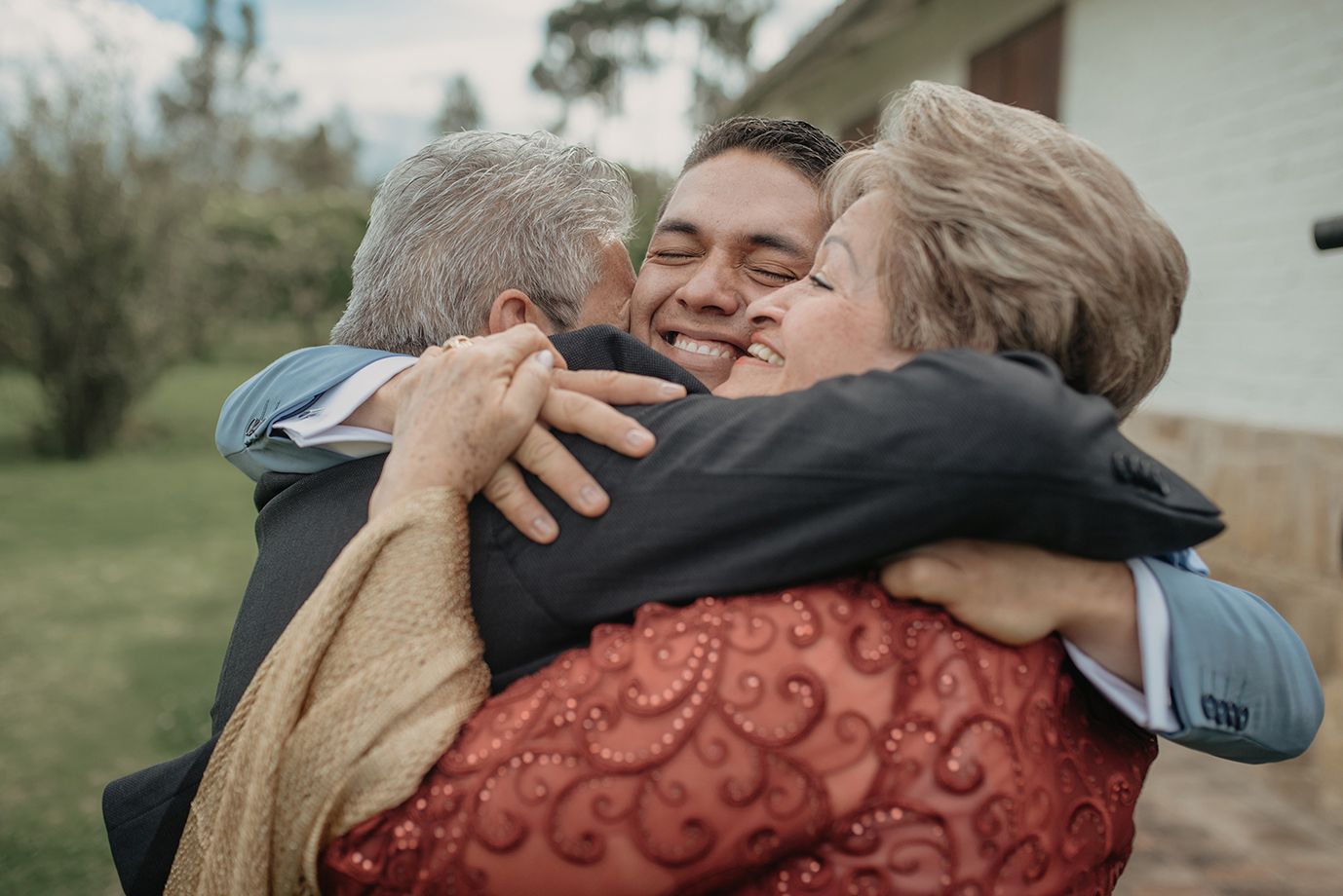 Groom sharing warm group hug with smiling elderly relatives at outdoor wedding celebration.