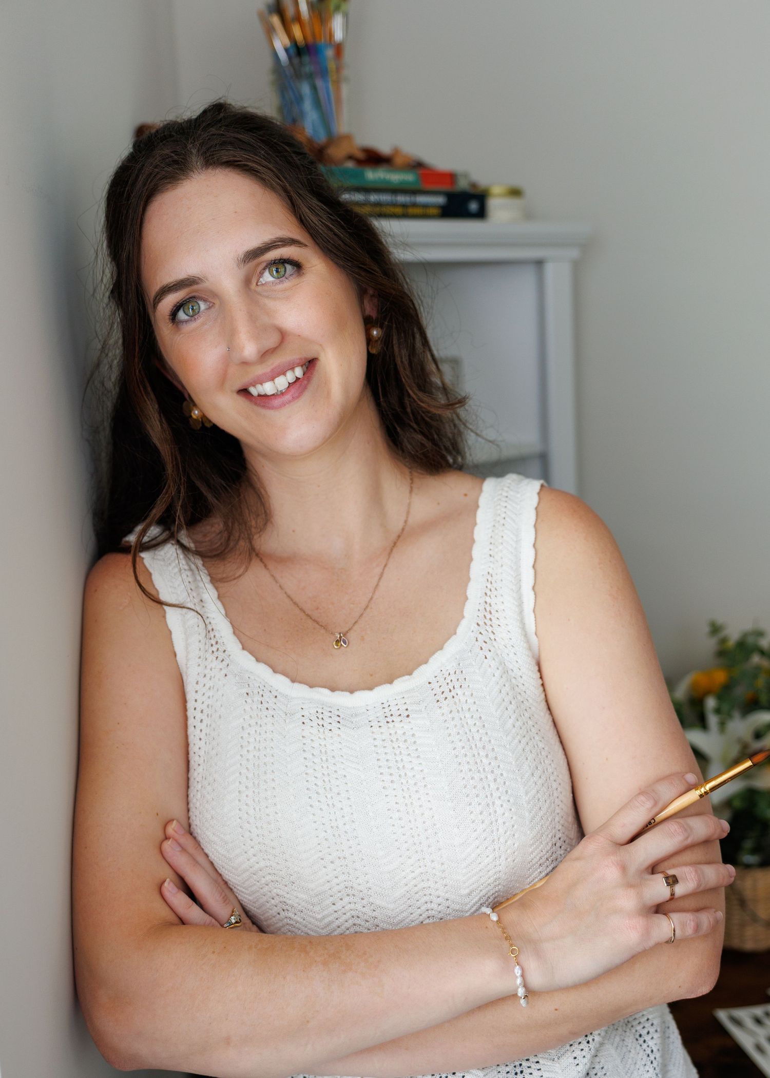 Smiling woman with brown hair holding a paintbrush, wearing a white knit top, standing in a bright art studio.