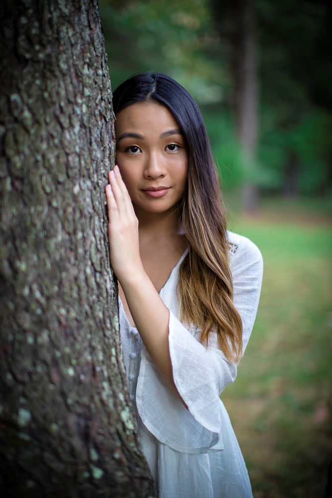 Young Asian woman in white dress leaning against a tree trunk in a lush green forest setting.