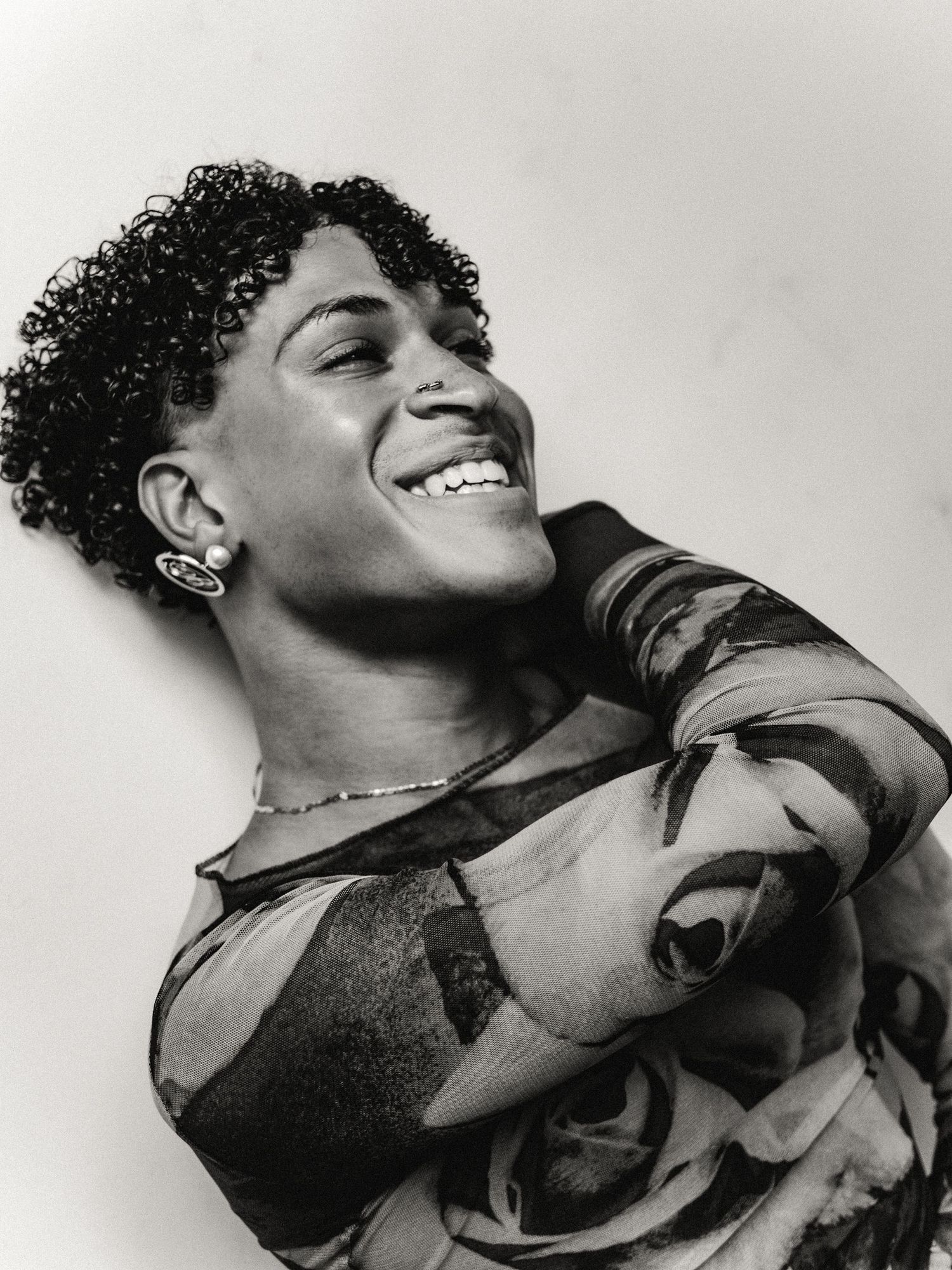Black and white portrait of a joyful person with curly hair, wearing a floral top, laughing with arms crossed.