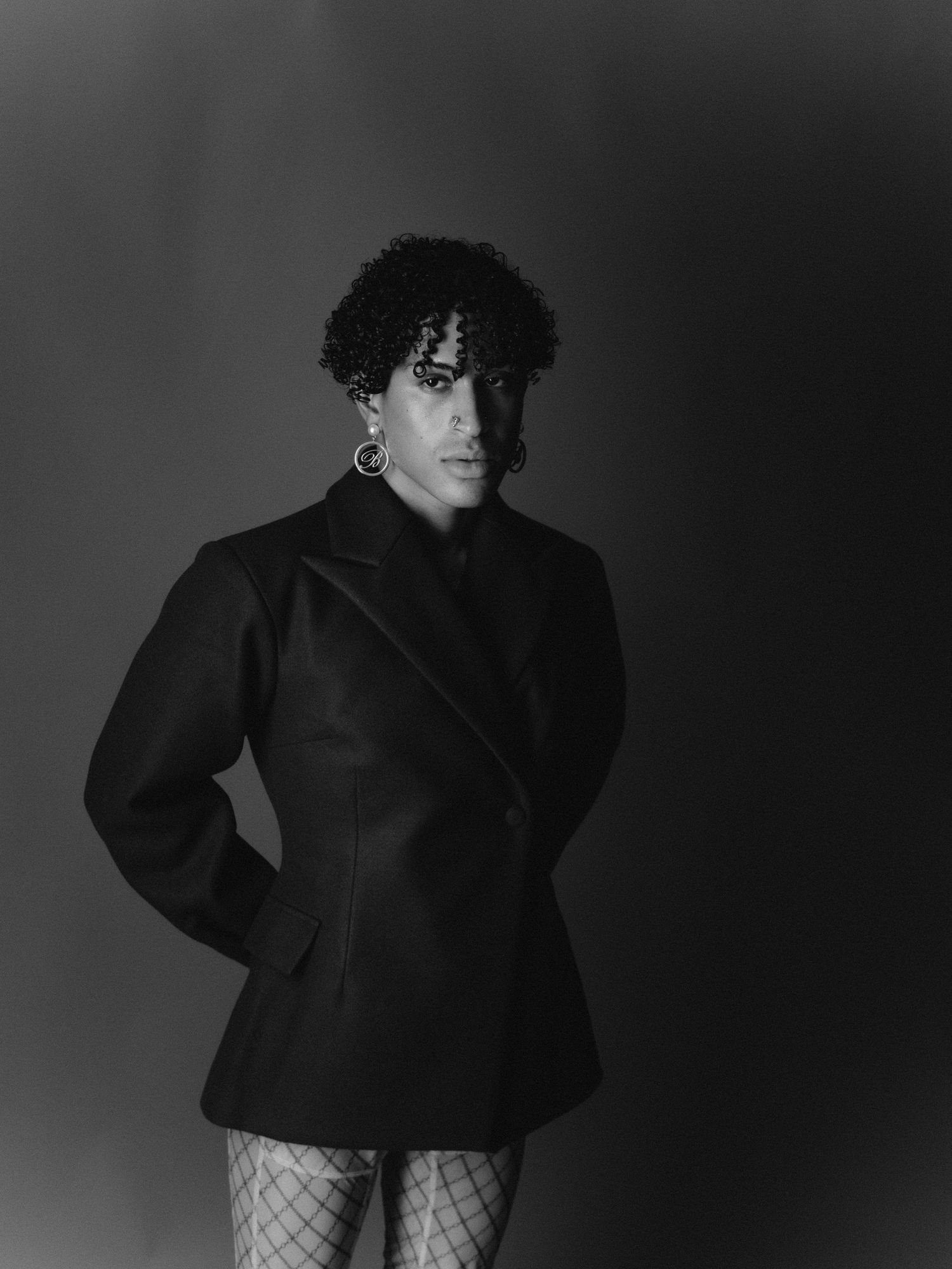 Black and white portrait of a person in oversized blazer, fishnet tights, and hoop earrings posing confidently.