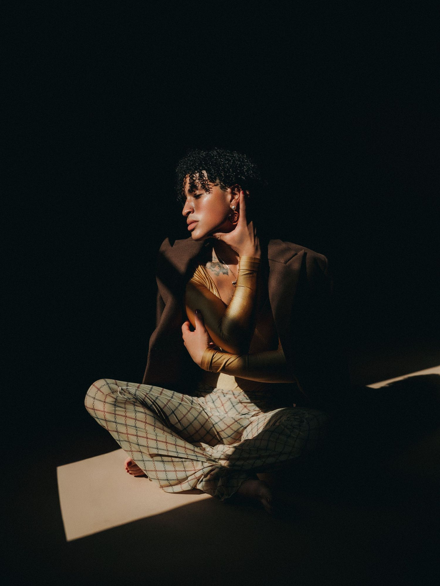 A person with curly hair sits cross-legged on the floor in dramatic lighting, wearing plaid pants and a dark blazer.