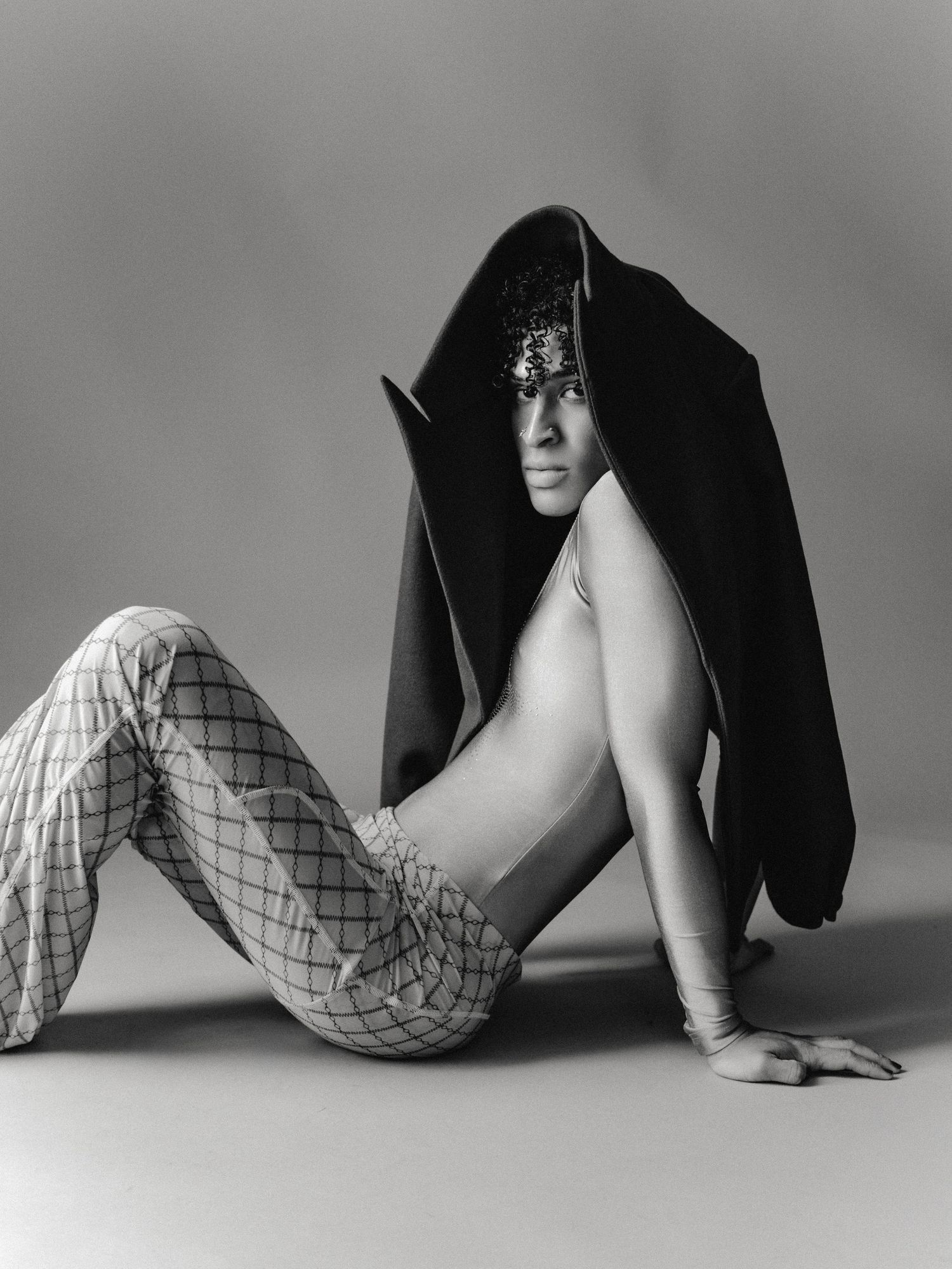 Black and white fashion photo of a model in plaid pants and dark draped fabric headpiece, seated on studio floor.