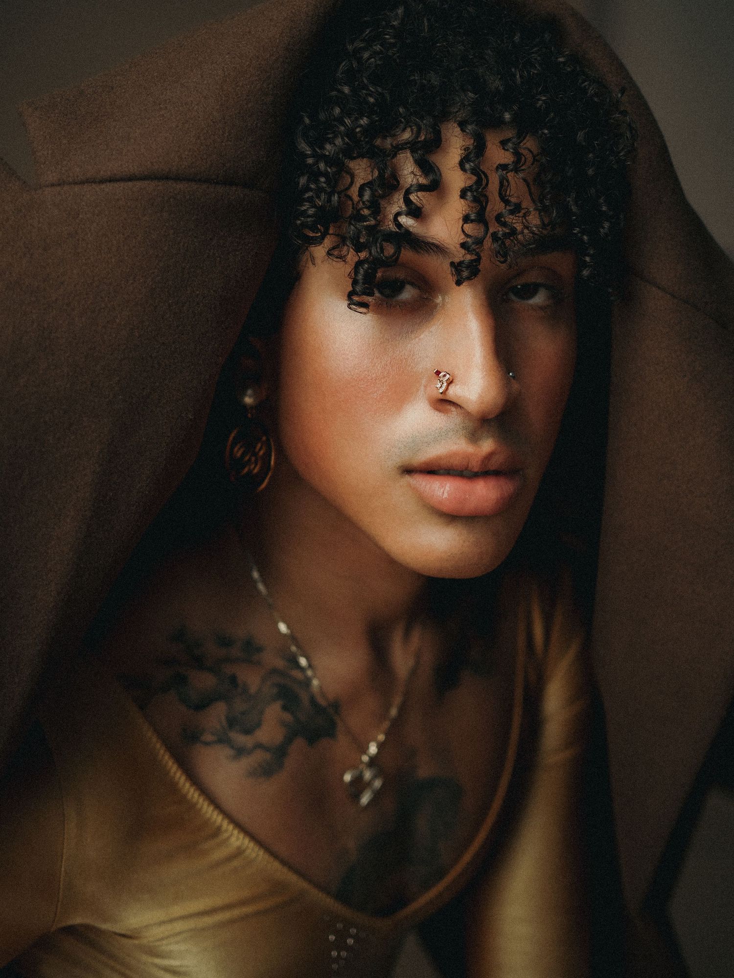 Close-up portrait of a person with curly hair, nose ring, hoop earrings, chest tattoos, and necklace draped in brown fabric.
