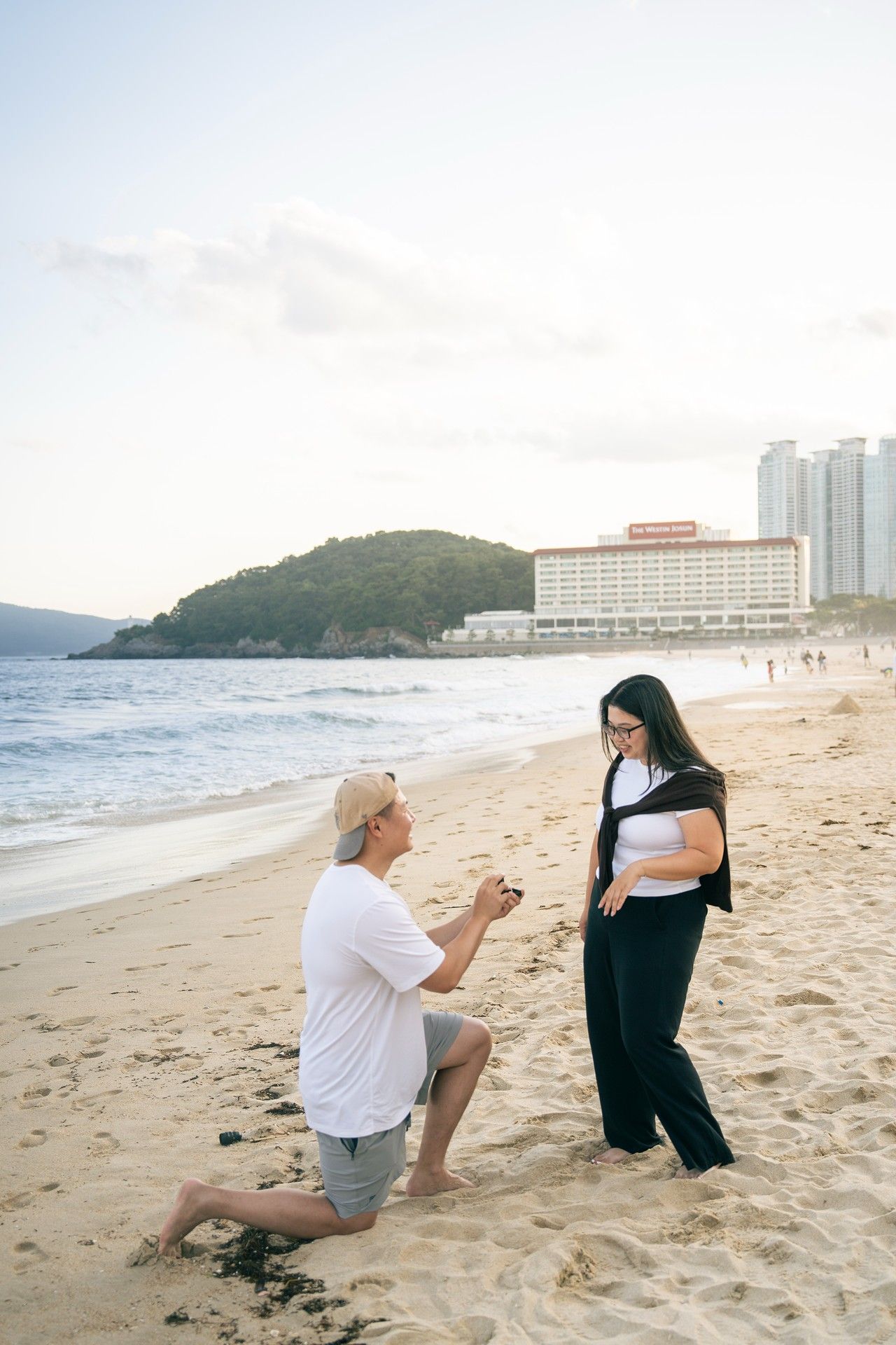 Man proposes on bended knee to woman on sandy beach with ocean waves and city buildings in background.