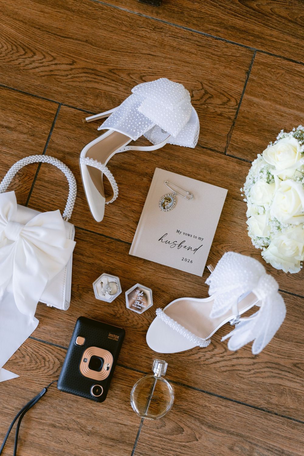 Bridal flat lay with white heels, pearl bag, Instax camera, perfume, ring boxes, vows card, and white floral bouquet on wood floor.