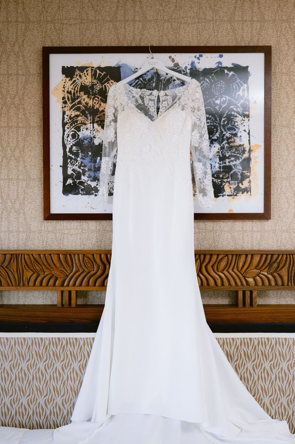 Elegant white lace long-sleeve wedding gown hanging on a hanger against a framed art piece in a hotel room.