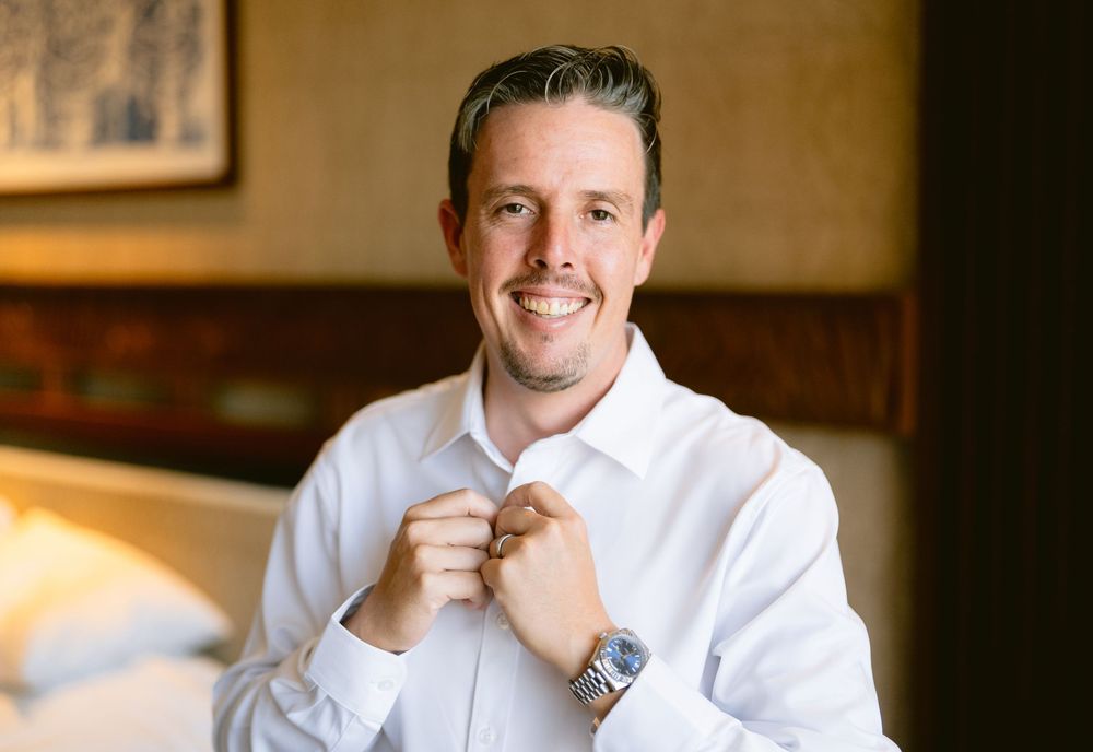 Smiling groom in white dress shirt adjusting his tie, wearing a watch, in an elegant hotel room.
