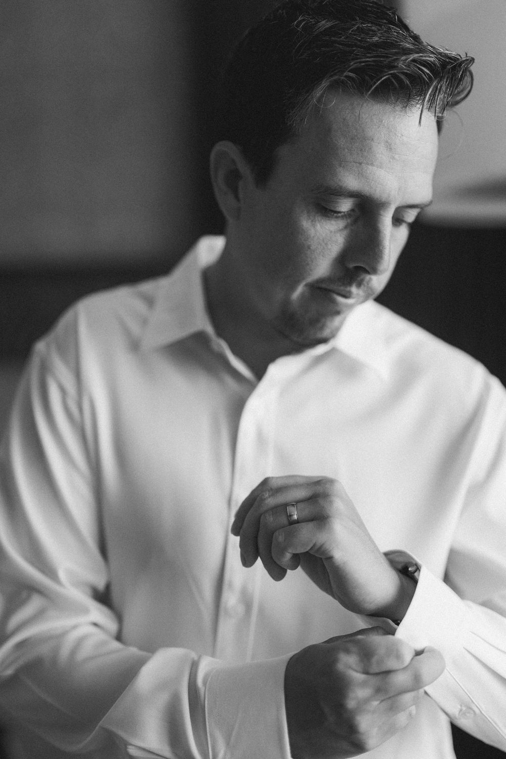 Black and white photo of a groom in a white dress shirt fastening his cufflinks before his wedding ceremony.