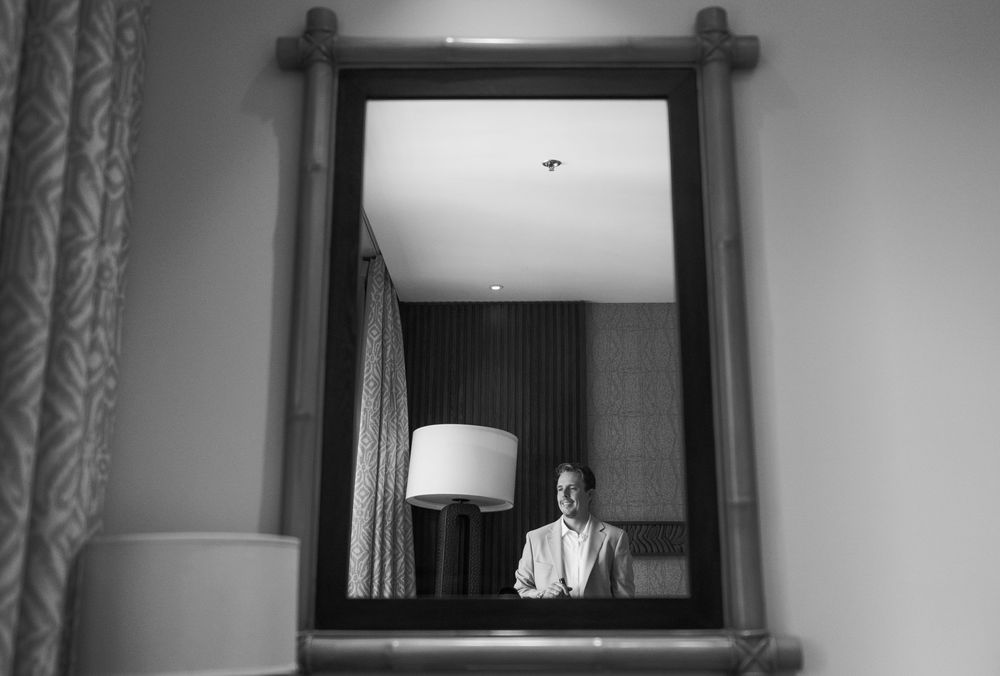 Black and white photo of a man in a suit reflected in an ornate mirror in an elegant hotel room.